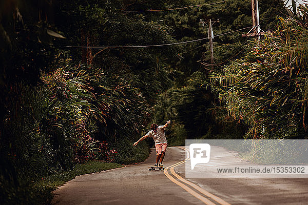 Mittelgroßer erwachsener männlicher Skateboarder  der auf einer Landstraße mit ausgestreckten Armen Skateboard fährt  Haiku  Hawaii  USA