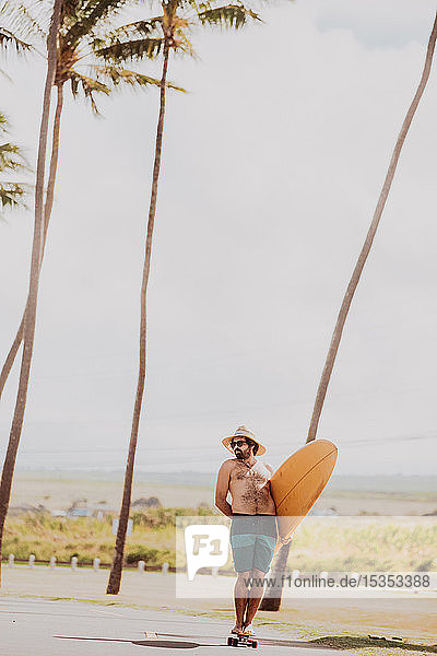 Mid adult male skateboarder carrying surfboard  standing on skateboard on coastal road  Haiku  Hawaii  USA