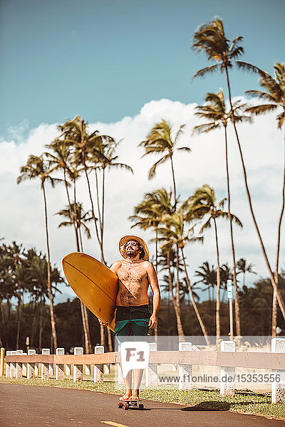 Mittelgroßer erwachsener männlicher Skateboarder mit Strohhut  steht auf Skateboard und trägt ein Surfbrett auf einer Landstraße  Portrait  Haiku  Hawaii  USA