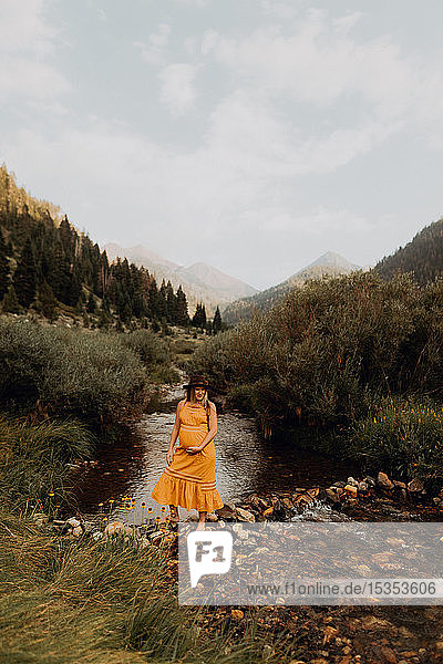 Schwangere Frau in orangefarbenem Maxikleid überquert Trittsteine im ländlichen Fluss  Mineral King  Kalifornien  USA