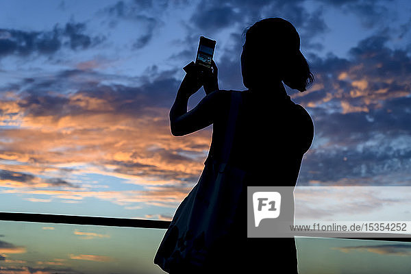 Silhouette einer Frau  die den Sonnenuntergang mit einem Smartphone fotografiert; Havanna  Kuba