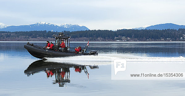Motorboot mit vier Personen in roten Jacken  die entlang einer Küstenlinie fahren  mit den Coast Mountains im Hintergrund  Blackie Spit  Crescent Beach; Surrey  British Columbia  Kanada