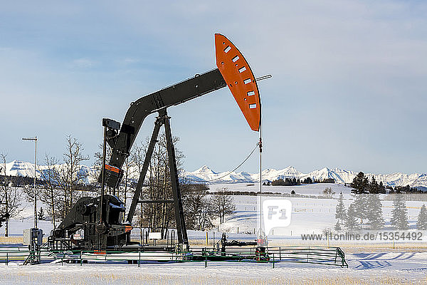 Pumpjack in a snow-covered field with snow-covered mountains and blue sky in the background  West of Airdrie; Alberta  Canada