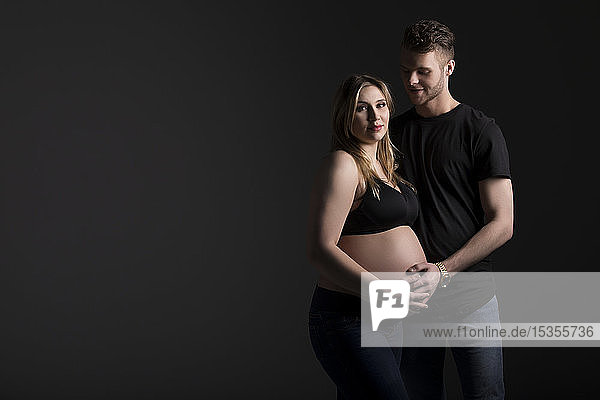 A young expectant couple with the mother looking at the camera while they are holding her belly in a studio on a black background: Edmonton  Alberta  Canada