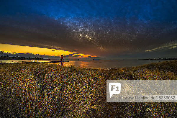 Leuchtturm Herd Groyne und dramatischer Sonnenuntergang mit leuchtenden Wolken und Strandgras im Vordergrund; South Shields  Tyne and Wear  England