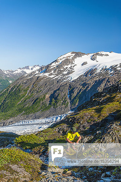 Ein Mann  der mit dem Rucksack unterwegs ist und seine Wasserflasche an einem kleinen Bach im Kenai Fjords National Park mit dem Exit Glacier im Hintergrund an einem sonnigen Sommertag in Süd-Zentral-Alaska auffüllt; Alaska  Vereinigte Staaten von Amerika
