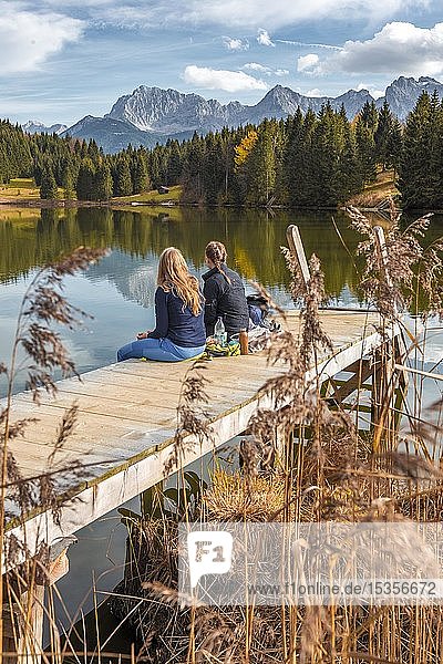 Zwei junge Frauen sitzen an einem Steg und essen eine Kleinigkeit  Geroldsee  Mittenwald  Karwendel  Bayern  Deutschland  Europa