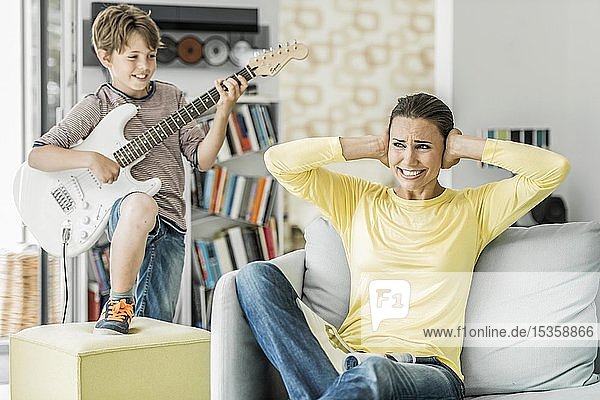 Mutter sitzt auf der Couch im Wohnzimmer und hält sich die Ohren zu  Sohn spielt E-Gitarre  Deutschland  Europa