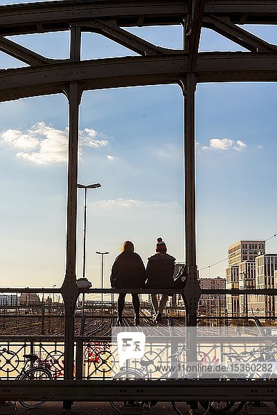 Zwei junge Frauen sitzen auf der Brüstung der Hackerbrücke über den Bahngleisen und schauen in die Ferne Gegenlicht München Oberbayern Bayern Deutschland Europa