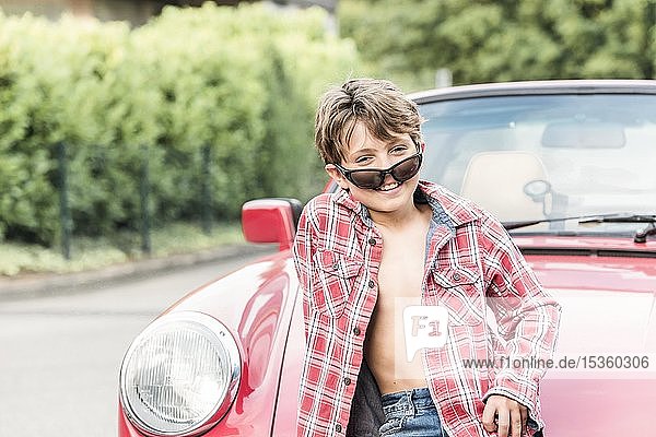 Junge  10 Jahre alt  mit Sonnenbrille und kariertem Hemd lehnt an einem roten Auto und schaut lächelnd in die Kamera  Deutschland  Europa
