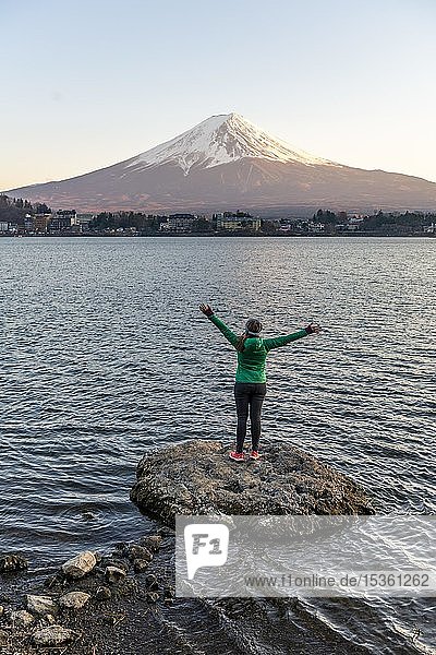 Abendstimmung  junge Frau steht auf einem Stein im Wasser und streckt die Arme in die Luft  Blick über den Kawaguchi-See  im Rücken der Vulkan Mt. Fuji  Präfektur Yamanashi  Japan  Asien