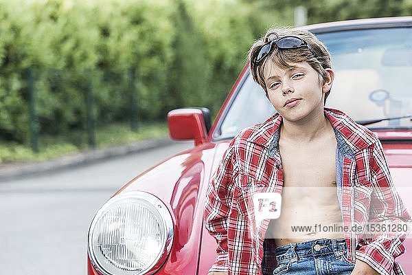 Junge  10 Jahre alt  mit Sonnenbrille und kariertem Hemd lehnt an einem roten Auto und schaut cool in die Kamera  Deutschland  Europa