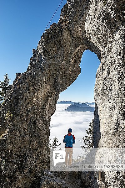 Young man stands in the rock gate Breitensteinfensterl with a view of the high fog above the valley  hiking trail to Breitenstein  Fischbachau  Bavaria  Germany  Europe