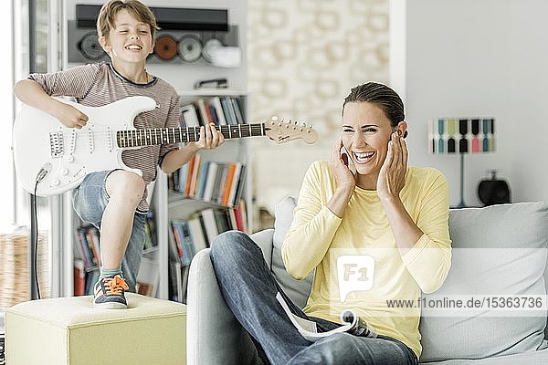 Mother sits laughing on the couch in the living room and covers her ears  son plays electric guitar  Germany  Europe