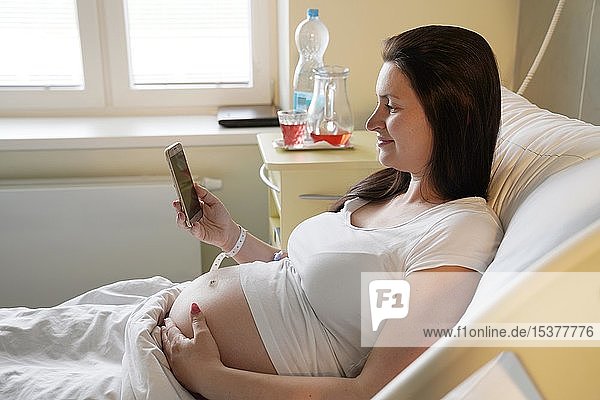 Risikoschwangerschaft  schwangere Frau liegt auf einem Entbindungsbett im Krankenhaus und schaut auf ein Mobiltelefon  Karlovy Vary  Tschechische Republik  Europa