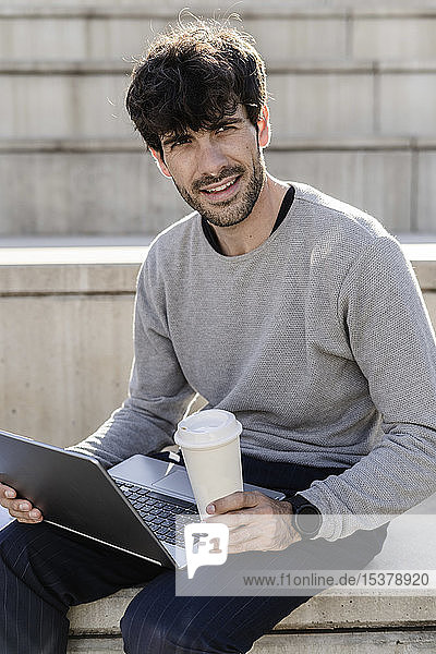 Mann sitzt auf einer Außentreppe mit Kaffee zum Mitnehmen und Laptop