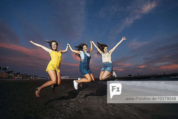Drei unbekümmerte Freundinnen springen bei Sonnenuntergang an den Strand