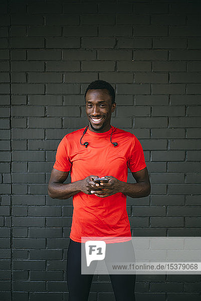 Portrait of a smiling young sportive man standing in front of a brick wall using his smartphone