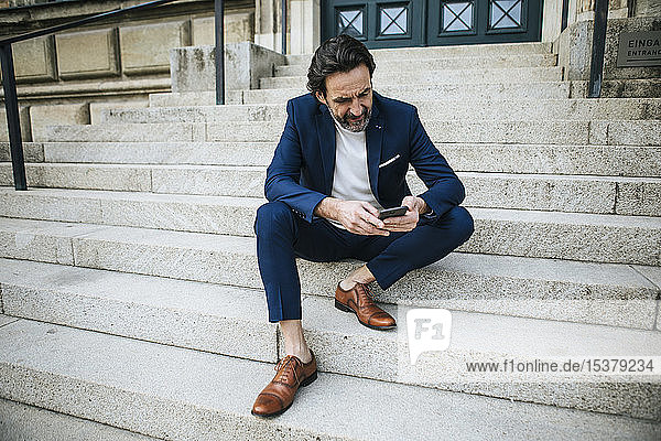 Bärtiger  reifer Geschäftsmann in blauem Anzug sitzt auf der Treppe und schaut auf sein Handy