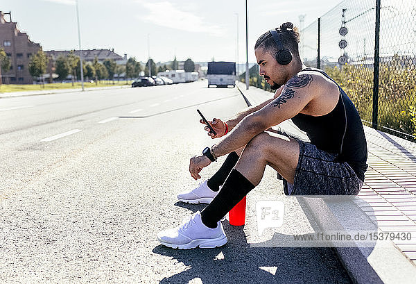 Sportlicher junger Mann mit Smartphone und Kopfhörern am Straßenrand sitzend