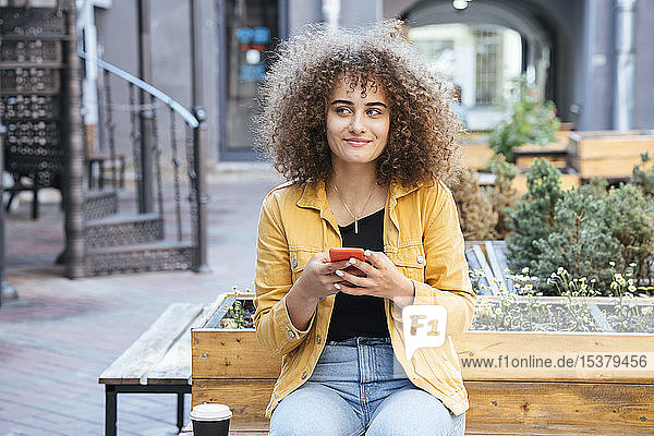 Portrait of smiling teenage girl sitting on bench with coffee and mobile phone