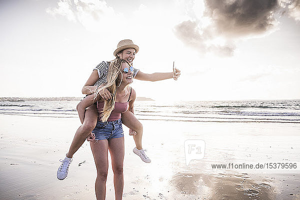Two girlfriends having fun on the beach  carrying each other piggyback  taking smartphone selfies