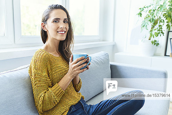 Porträt einer jungen Frau  die Kaffee trinkt  auf dem Sofa sitzt und in die Kamera lächelt