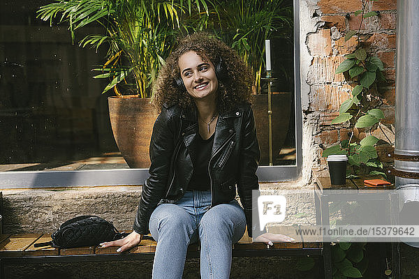 Portrait of smiling teenage girl sitting at outdoor cafe listening music with headphones and smartphone