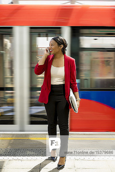 Porträt einer lachenden Geschäftsfrau am Telefon auf dem Bahnsteig stehend  London  UK