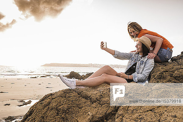Zwei Freundinnen sitzen am felsigen Strand und nehmen sich ein Smartphone
