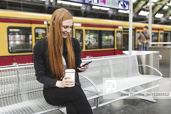 Rothaarige junge Frau mit Kaffee zum Warten am Bahnsteig per Smartphone  Berlin  Deutschland