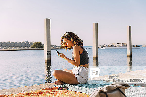 Junge Frau sitzt auf einem Pier am Meer und benutzt ein Smartphone