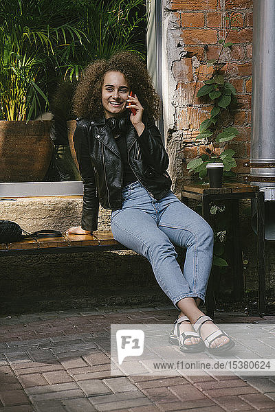 Porträt einer glücklichen Teenagerin am Telefon  die in einem Café im Freien mit Kaffee zum Mitnehmen sitzt