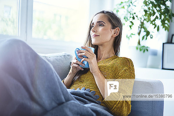 Beautiful young woman enjoying cup of coffee while lying on sofa under blanket