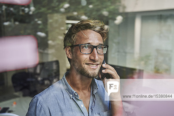 Casual young businessman on the phone behind windowpane in office