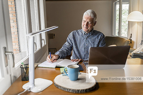 Älterer Mann sitzt zu Hause vor dem Laptop und macht Notizen im Notizbuch