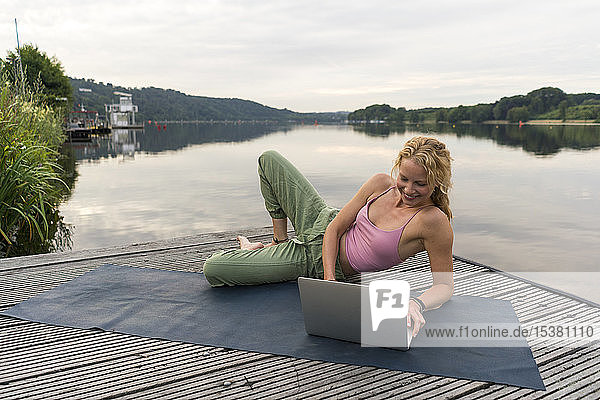 Smiling young woman using laptop on a jetty at a lake
