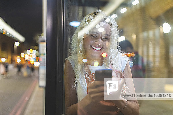 Young woman in London at night looking at her smartphone and waiting for the bus