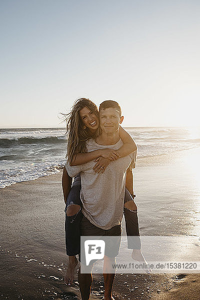 Portrait of happy young man carrying girlfriend piggyback on the beach at sunset