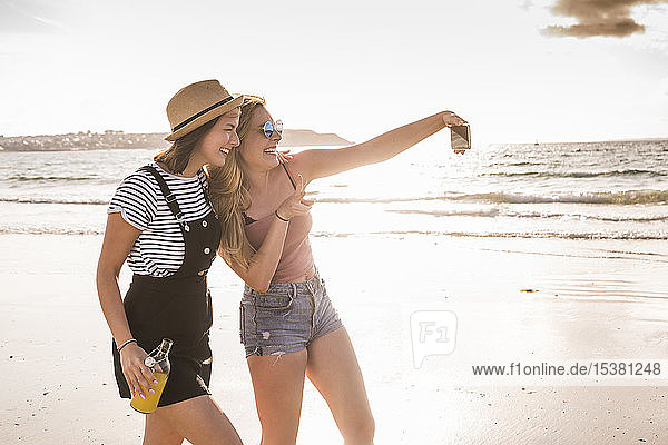Zwei Freundinnen amüsieren sich  gehen am Strand spazieren  nehmen selbst ein Smartphone