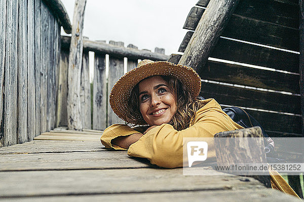 Junge lächelnde Frau mit gelbem Mantel und Hut auf einer Holztreppe  seitwärts blickend
