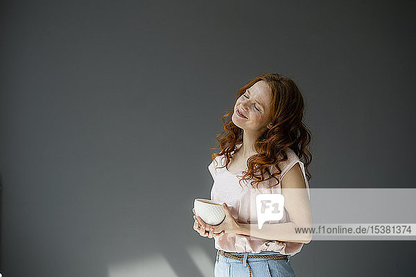 Porträt einer glücklichen rothaarigen Frau mit Kaffeeschüssel vor grauem Hintergrund