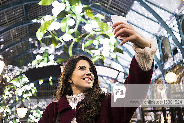 Porträt einer lächelnden jungen Frau  die sich im beleuchteten Pavillon mit einem Smartphone selbstständig macht