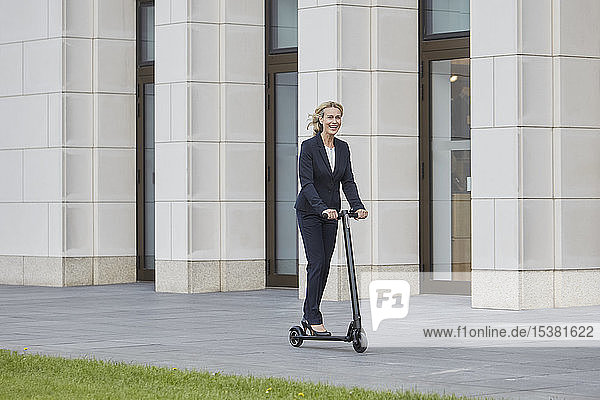Geschäftsfrau auf E-Scooter fährt an Bürogebäude in der Stadt vorbei