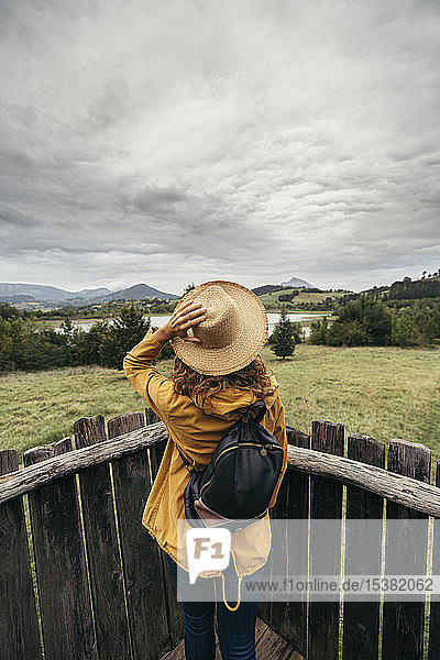 Junge Frau in gelbem Mantel und Rucksack mit einer Hand  die einen Hut im Kopf hält und auf einem Holzbalkon die Seelandschaft betrachtet
