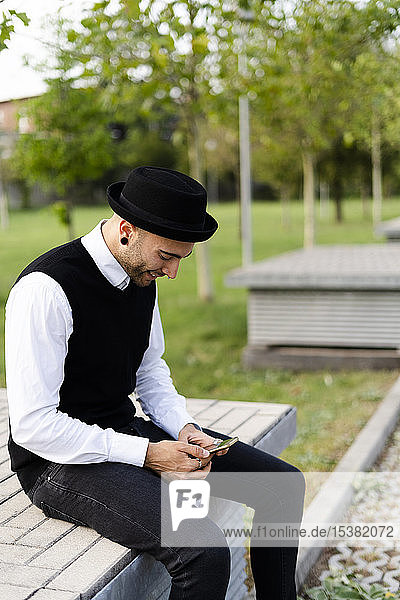 Stylish man sitting on bench outdoors looking at cell phone