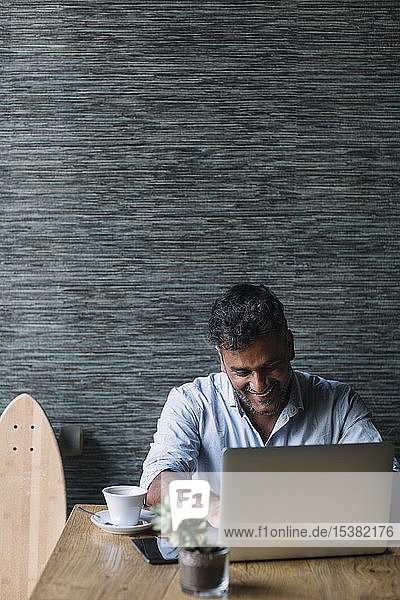 Lächelnder Gelegenheits-Geschäftsmann mit Skateboard mit Laptop in einem Cafe