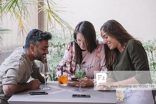 Lächelnde Freunde sitzen mit Getränken am Tisch und schauen auf das Smartphone