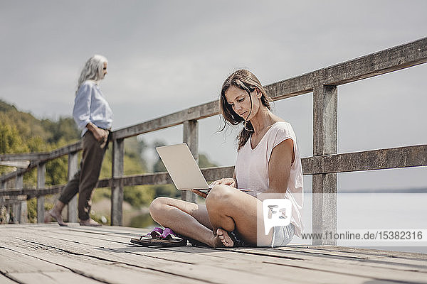 Frau auf Holzbrücke sitzend  mit Laptop  Mutter im Hintergrund stehend