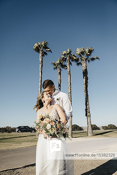 Glückliche Braut und Bräutigam umarmen sich an Palmen unter blauem Himmel